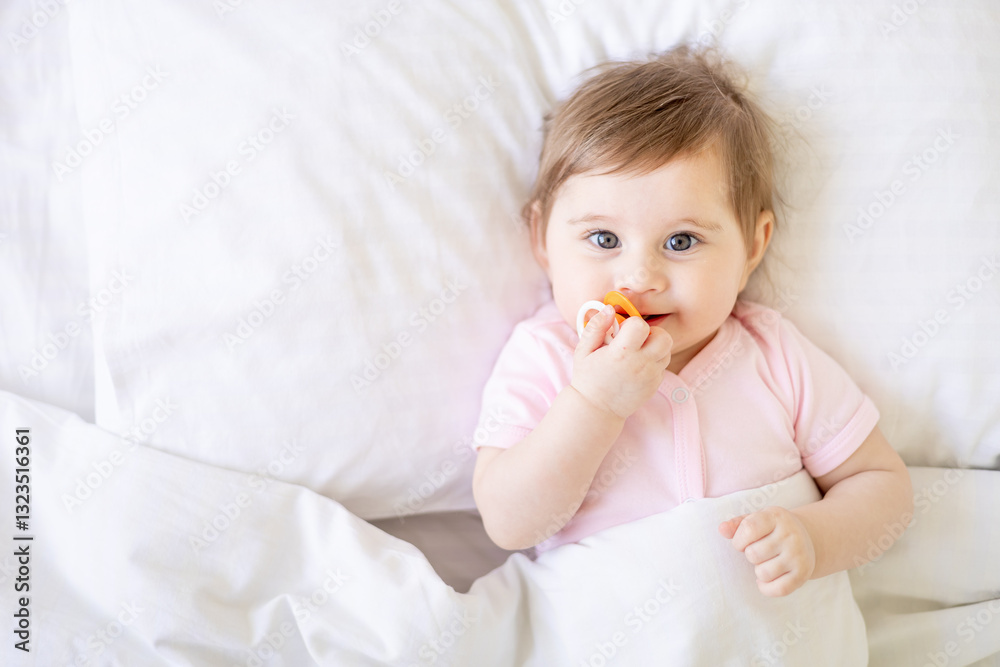 baby sucking a pacifier lying on a white cotton bed under a blanket on the bed at home and smiling, cute little baby girl in a pink bodysuit, close-up baby sucking a pacifier lying on a white cotton bed under a blanket on the bed at home and smiling, cute little baby girl in a pink bodysuit, close-up