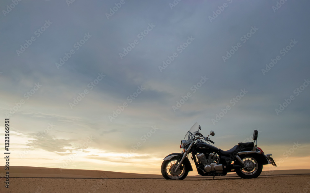 Fototapeta premium A lone motorcycle stands on the sandy surface of the Namib Desert against a dramatic cloudy sky. The sunset lighting highlights the contrast between nature and machine.