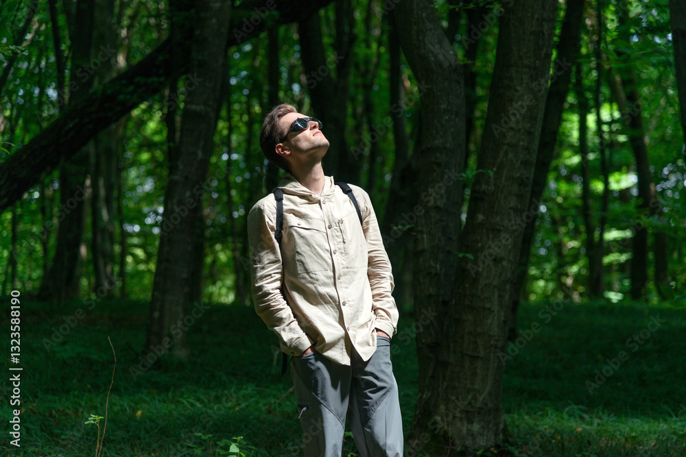 Adventurous man hiking in a forested mountain area, looking up while standing in the dark forest. Sunlight filters through the lush green trees. Adventurous man hiking in a forested mountain area, looking up while standing in the dark forest. Sunlight filters through the lush green trees.