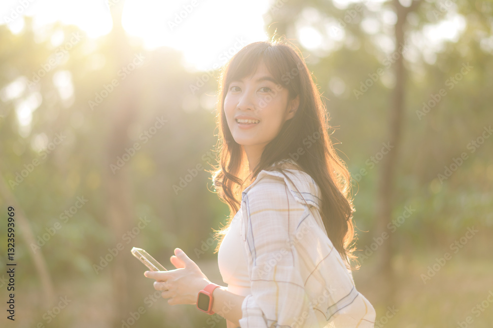 Naklejka premium Asian woman enjoying summer sunlight while breathing fresh air in serene outdoor setting, holding smartphone with cheerful expression