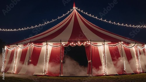 red white striped circus tent at night, illuminated by lights and featuring large banners on the sides. The top of the tent is decorated with red stripes and white canvas material. 