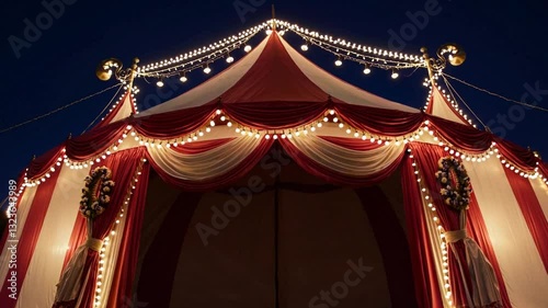 red white striped circus tent at night, illuminated by lights and featuring large banners on the sides. The top of the tent is decorated with red stripes and white canvas material. 