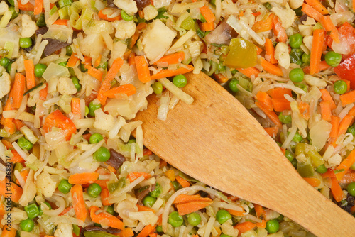 A close-up, top-down view of stir-fried mixed vegetables in a pan, with a wooden spoon resting on top, highlighting the colorful ingredients and cooking process