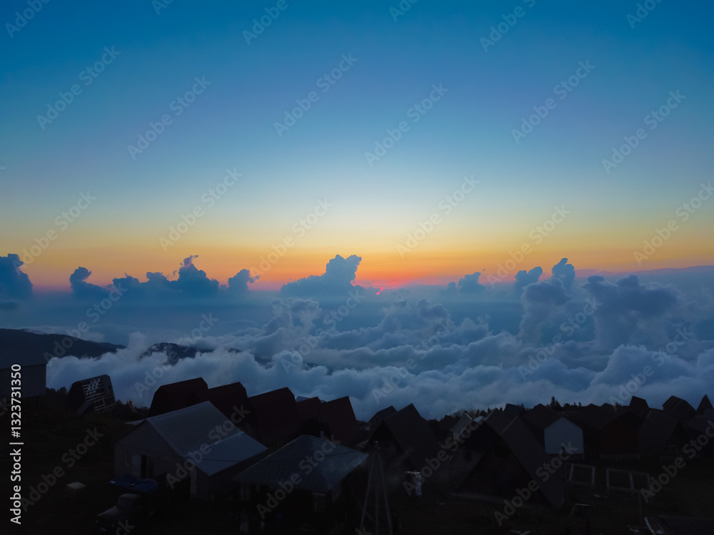 Sun sets over the mountain village, illuminating the sea of clouds below and creating a striking contrast between the dark silhouettes of the village and the bright sky