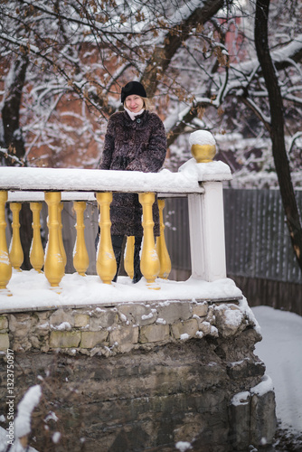 Obraz na płótnie A woman in a karakul fur coat stands by the railing of a historic house, surrounded by the charm of a snowy winter scene. w salonie