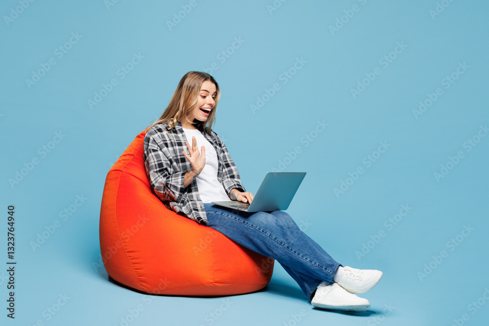 Fototapeta premium Full body young IT woman she wear checkered grey shirt casual clothes sit in bag chair hold use work on laptop pc computer waving hand isolated on plain pastel blue cyan background. Lifestyle concept