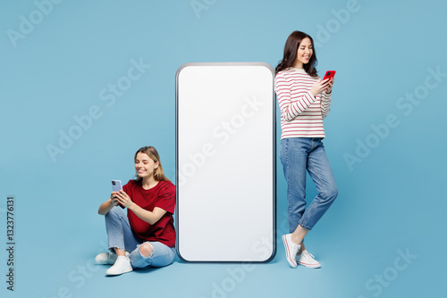 Full body young happy couple two friends women wearing red casual clothes together stand sit near big huge blank screen mobile cell phone with area using smartphone isolated on plain blue background.