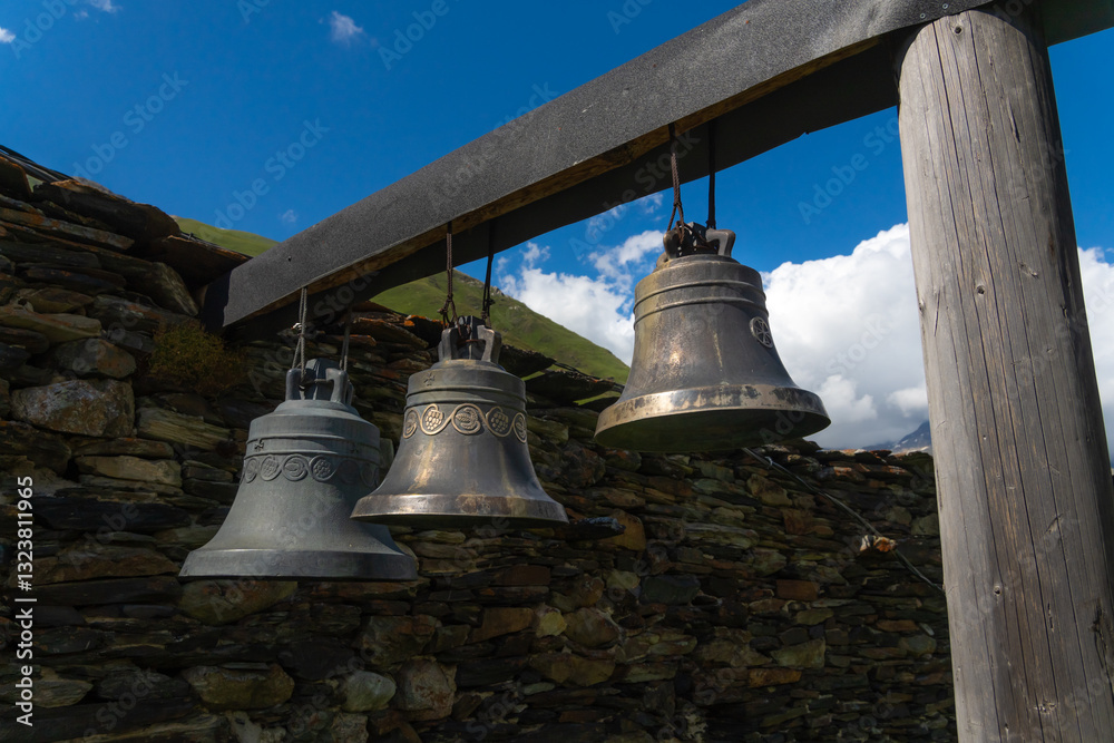Three large bells hang from a wooden beam in front of a stone wall, with green mountains and a blue sky in the background, creating a serene and traditional scene Three large bells hang from a wooden beam in front of a stone wall, with green mountains and a blue sky in the background, creating a serene and traditional scene