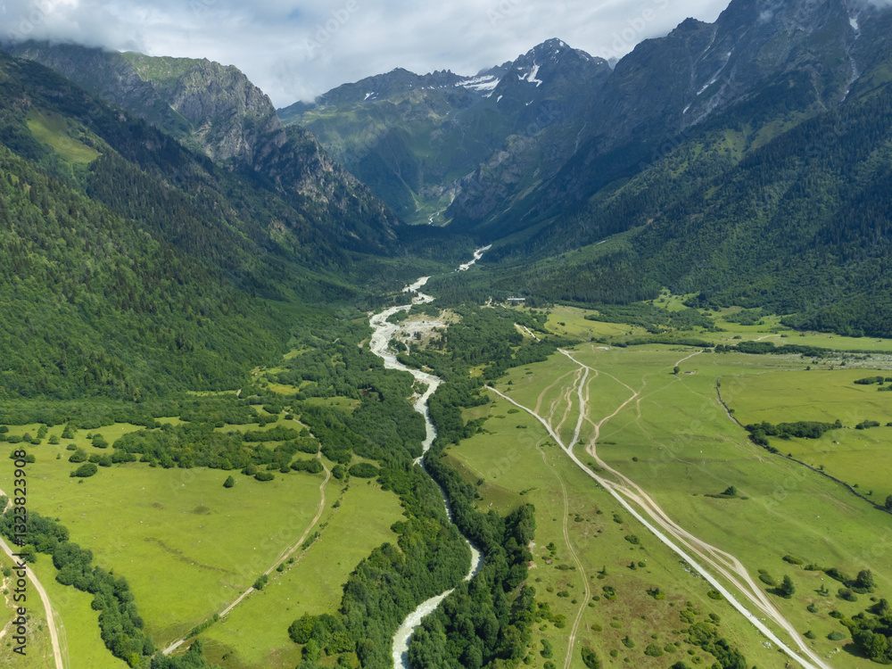 Awe-inspiring aerial perspective captures a pristine river meandering through a vibrant green valley, nestled amidst a breathtaking mountain range, creating a picturesque landscape