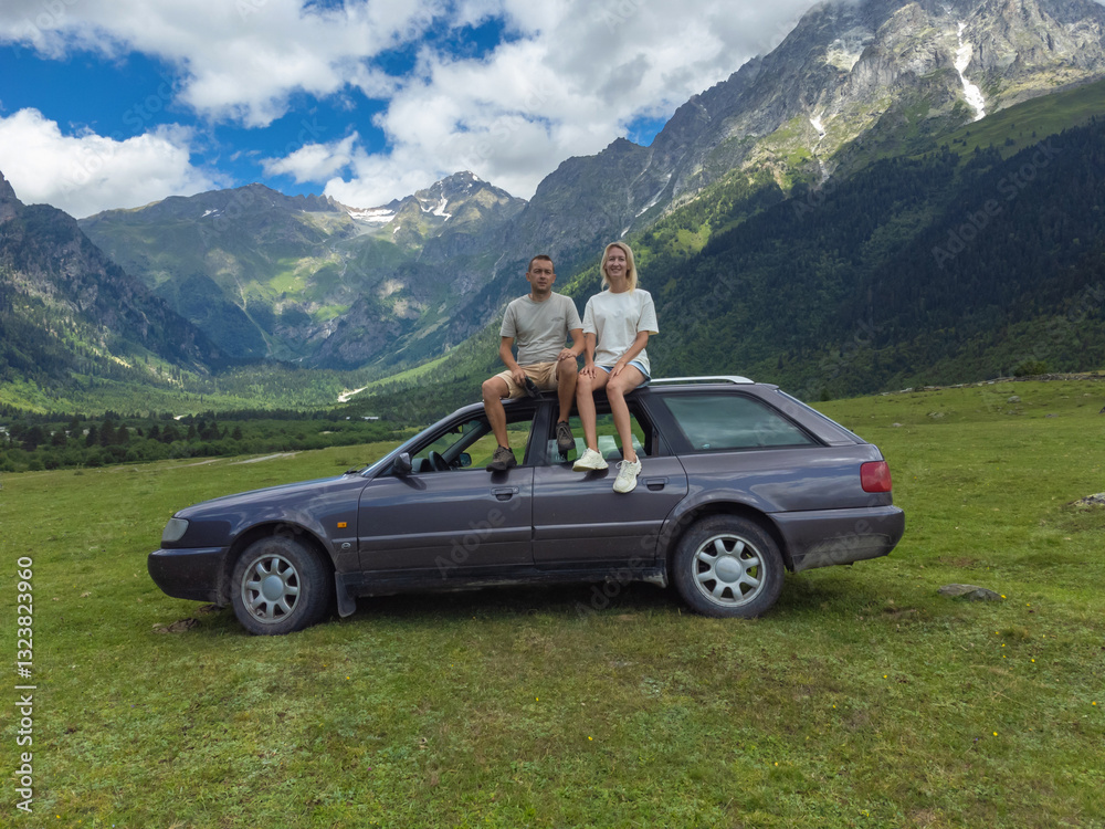 Young couple sitting on the roof of their car, taking in the breathtaking panoramic view of a majestic mountain range during a memorable road trip