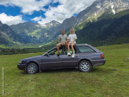 Obraz na płótnie Young couple sitting on the roof of their car, taking in the breathtaking panoramic view of a majestic mountain range during a memorable road trip w salonie