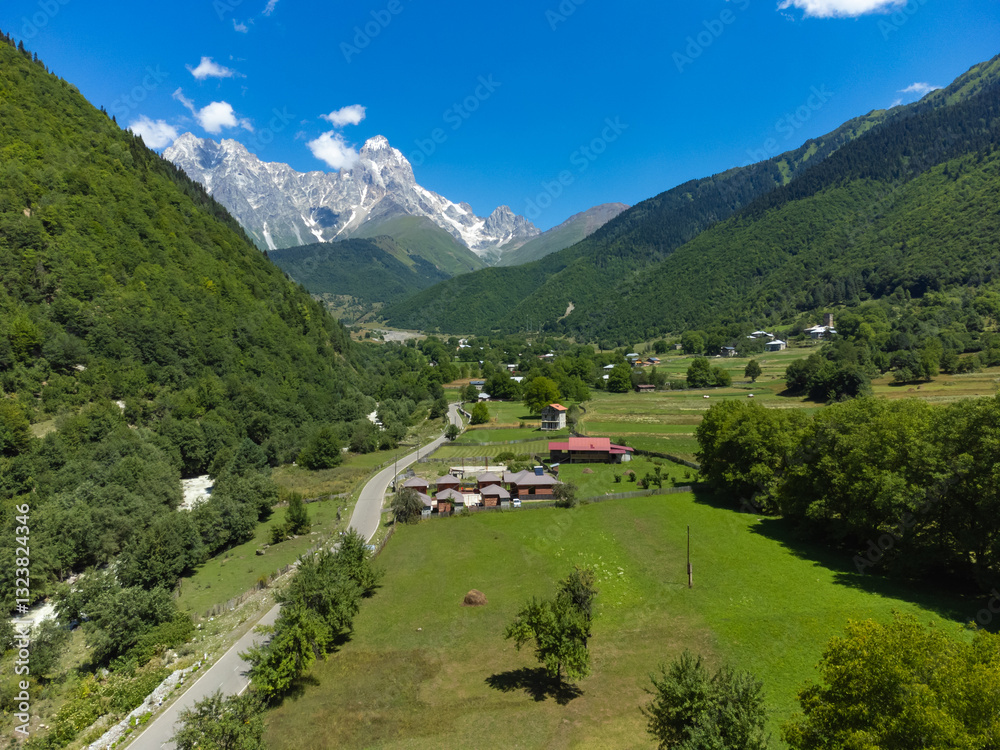 Road crossing a green valley leading to snow-capped mount ushba, dominating the landscape of svaneti region in georgia, under a bright blue sky with white clouds