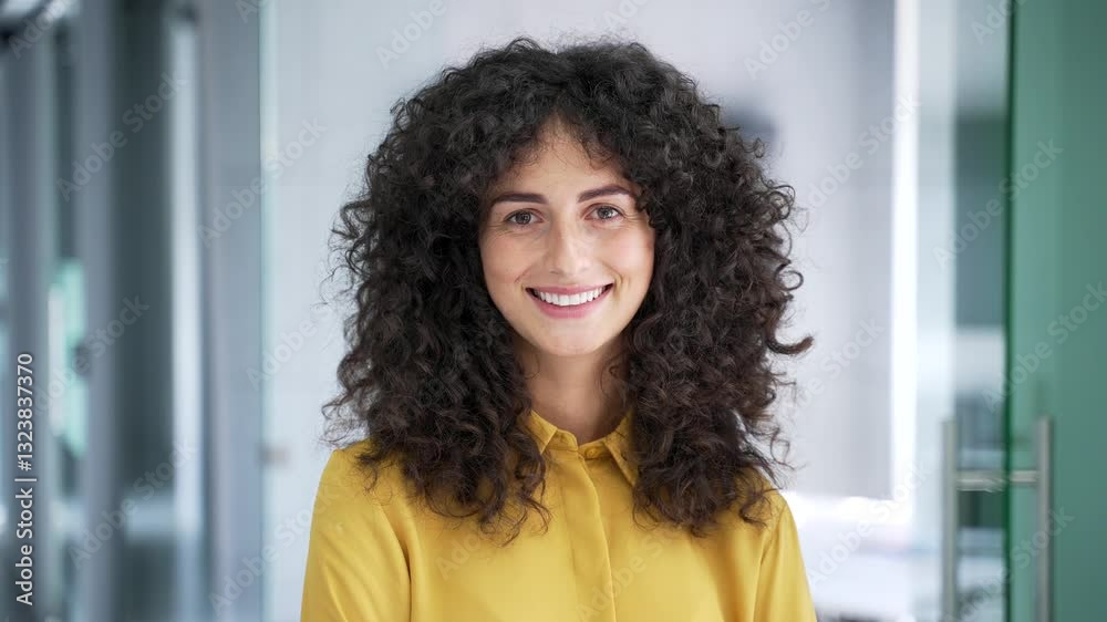 Portrait of young smiling businesswoman standing in a modern business office. Positive happy curly brunette woman in yellow shirt is posing looking at camera. Headshot of successful manager. Close up Portrait of young smiling businesswoman standing in a modern business office. Positive happy curly brunette woman in yellow shirt is posing looking at camera. Headshot of successful manager. Close up