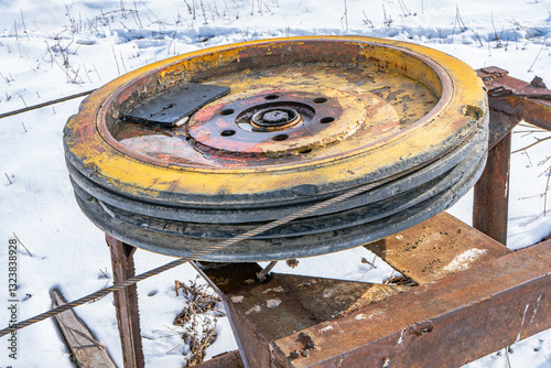 A large metal pulley wheel, worn with patches of yellow paint, rests on a rusted frame in a snow-covered field. This relic of industrial machinery stands out against the pristine white backdrop photo