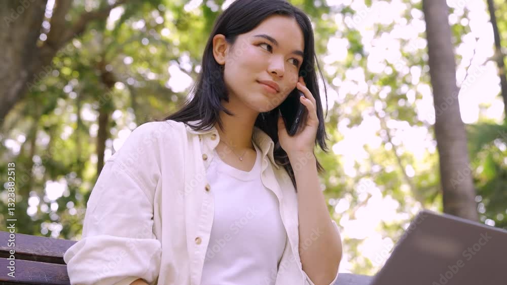 Young Japanese woman teleworking on her laptop and talking on the phone while sitting on a bench in a park Young Japanese woman teleworking on her laptop and talking on the phone while sitting on a bench in a park