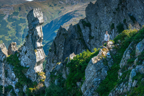 Fototapete Male climber standing on rocky peak surrounded by mountain cliffs and distant vi