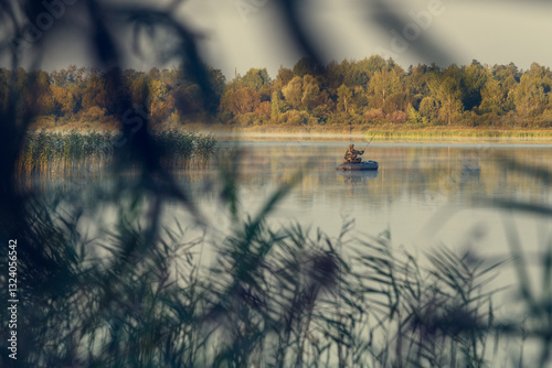 A fisherman in a rubber boat on a calm lake early in the morning, seen from afar through the reeds.