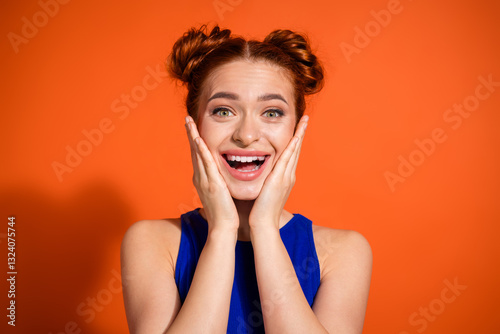 Joyful young woman with playful hairstyle posing in casual blue top against bright orange studio background expressing happiness