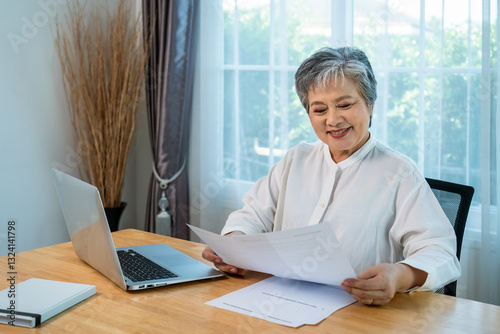 Senior Asian businesswoman manager smiling while reading documents at office desk with laptop