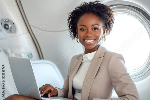 Portrait of smiling african american businesswoman in light business suit sitting in airplane cabin with laptop on her lap