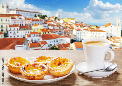 Cup of coffee and plate of traditional portuguese pastries Pastel de nata on wooden table over Alfama district background. Lisbon, Portugal