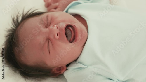 Close-up of newborn baby crying while lying on bed. The infant, dressed in a light blue outfit, appears distressed, showcasing natural emotions and early childhood expressions.