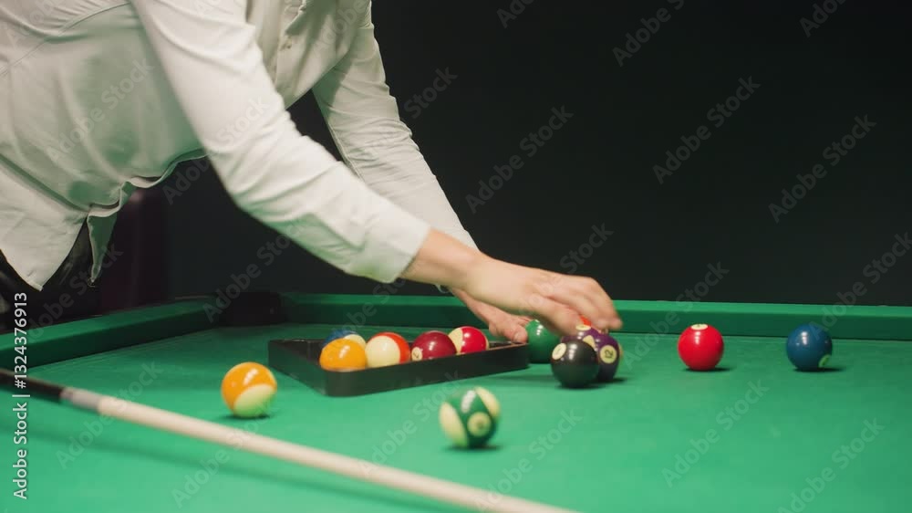 Person in white shirt using triangle rack to arrange billiard balls on green pool table. Cue stick lying nearby, balls scattered before being set for break shot. Dark background