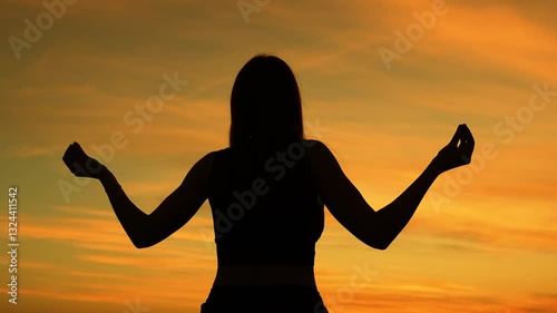 Silhouette of calm woman meditating at sunset dark outdoors. Mindful woman does spiritual practice to find harmony in evening park. Woman practicing yoga asana silhouette under sunset cloudy sky