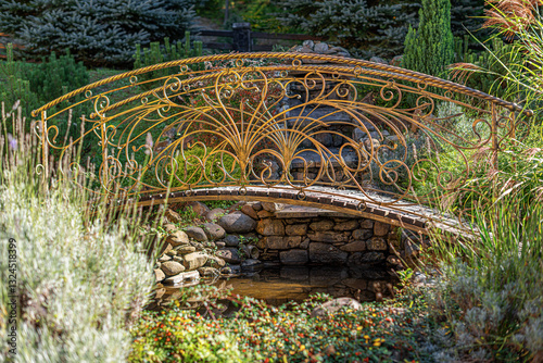Obraz na płótnie A beautifully crafted wrought iron bridge with intricate golden detailing spans a small garden stream, surrounded by lush greenery and bathed in warm sunlight. w salonie