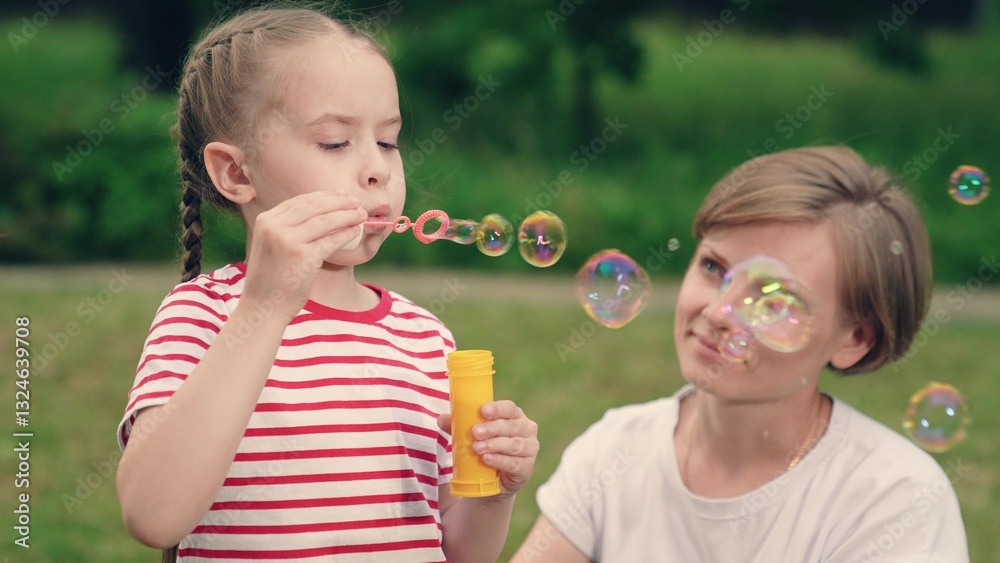 Happy fun kid, girl, mother blows soap bubbles against background of green trees. Child play outdoor concept. Daughter, mom play with soap bubbles in park summer day. Happy family relaxing in nature Happy fun kid, girl, mother blows soap bubbles against background of green trees. Child play outdoor concept. Daughter, mom play with soap bubbles in park summer day. Happy family relaxing in nature