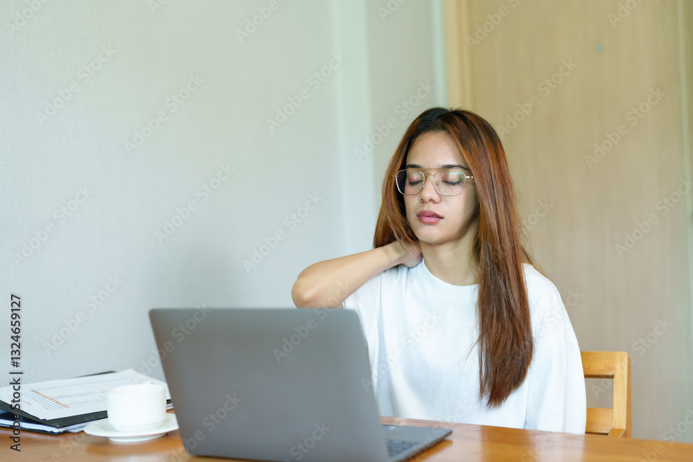 Asian young woman wearing glasses, stretching her neck while working on a laptop at home. Feeling tired from long screen time, dressed in a white t-shirt, sitting at a wooden table. Asian young woman wearing glasses, stretching her neck while working on a laptop at home. Feeling tired from long screen time, dressed in a white t-shirt, sitting at a wooden table.
