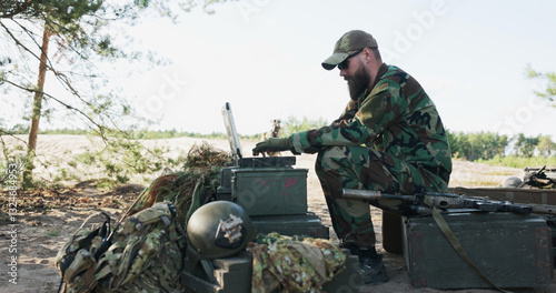 The army commander sits behind a computer in the field, creating reports for the base, as a soldier approaches with a map. They discuss details while soldiers guard the camp in the background. photo