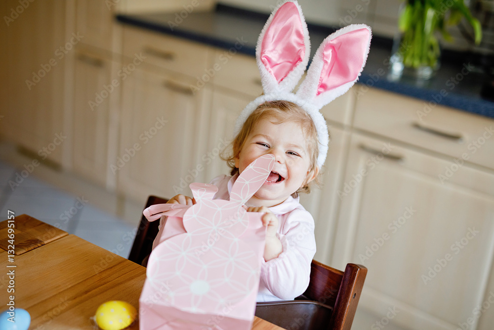 Cute little toddler girl wearing Easter bunny ears playing with colored pastel eggs. Happy baby child unpacking gifts. Adorable healthy smiling kid in pink clothes enjoying family holiday