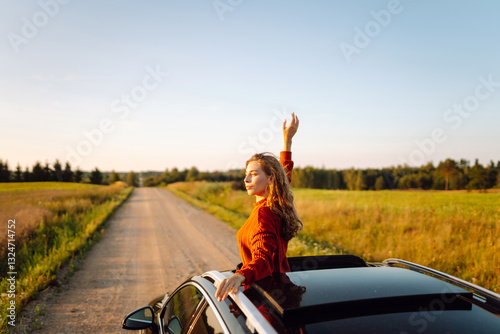 Young woman feels freedom leaning out of car window. Beautiful female traveler enjoying sunset from car. Car travel, leisure concept. Active lifestyle.