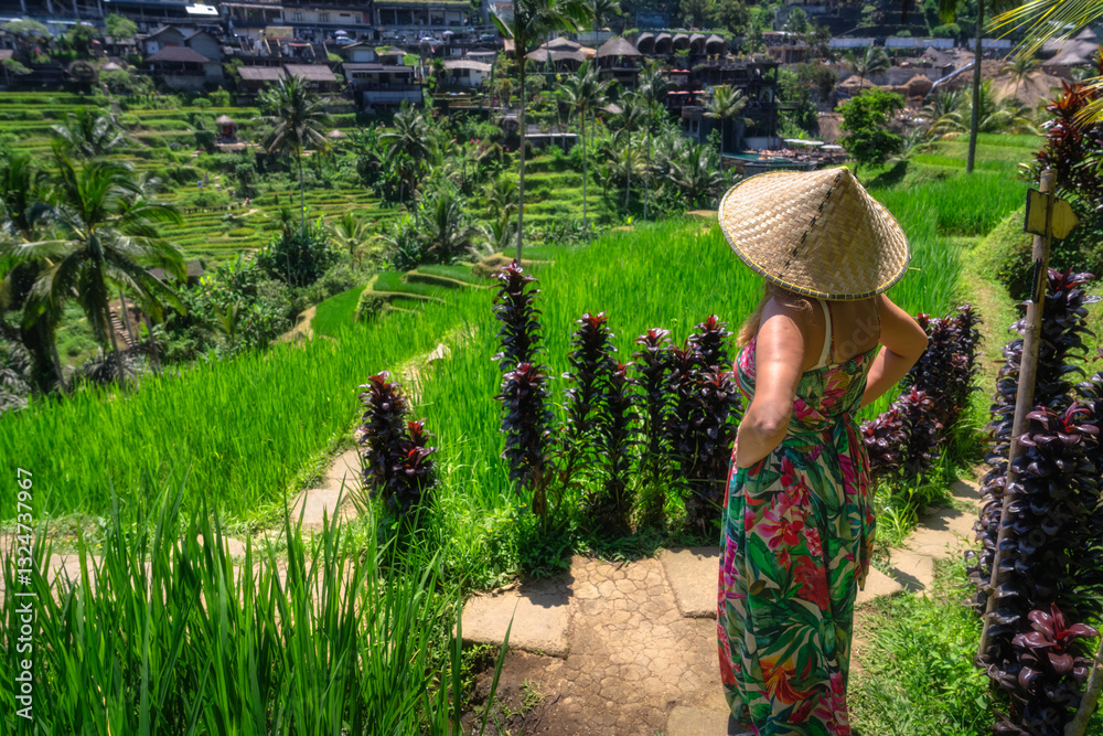 A woman, dressed in elegant attire and straw hat, stands gracefully amidst lush green rice terraces, fully immersing herself in mesmerizing tropical scenery that surrounds her, Ubud, Bali, Indonesia