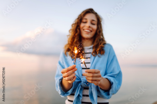Beautiful woman with curly hair having fun on beach holding sparklers in hands. Young tourist enjoying sunset outdoors. Freedom, enjoyment, holiday concept. photo
