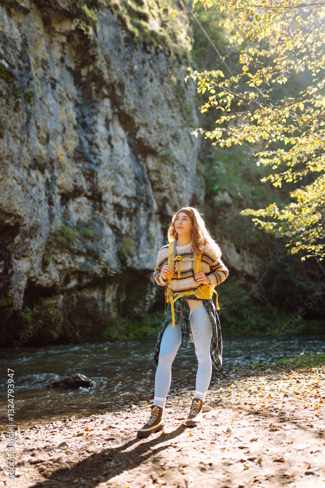 Naklejka premium Young woman with a hiking yellow backpack hiking in the mountains near a mountain river. Happy female traveler enjoying sunny weather outdoors. Travel concept, hiking. Active lifestyle.