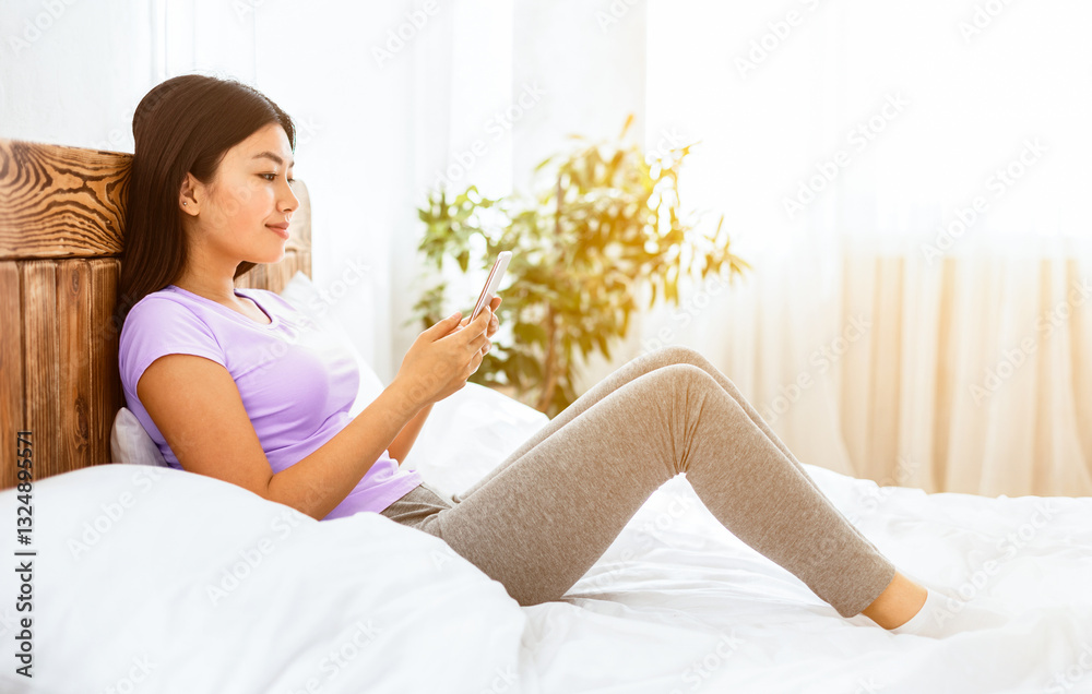 Japanese Girl Using Cellphone Texting In Chat Sitting In Bed In Bedroom On Weekend. Mobile App Concept