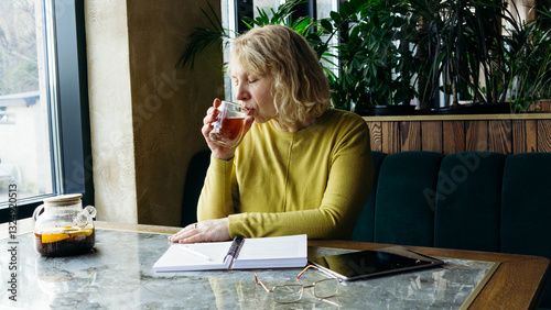 A senior businesswoman enjoys a beverage while using a tablet, embracing the freelance remote work concept. Her relaxed demeanor in a modern workspace