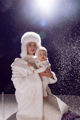 A mother hugs her daughter in her arms in white fur hats on Christmas Day. Snow is falling, the counterlight is in the back