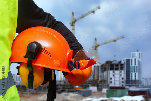 Construction worker in a high-visibility vest holding an orange safety helmet and protective gear at a building site.