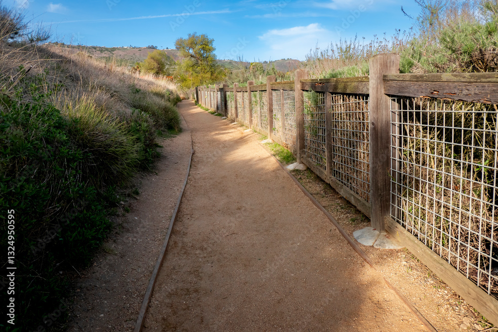 Dirt trail leading to the mountains up a slow and steady climb uphill, surrounded with well manicured greenery and wood post fence, blue skies with clouds