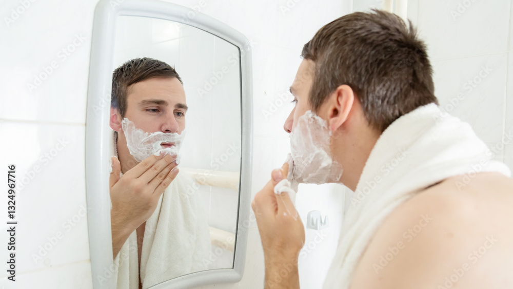 Young man with naked torso shaving his beard with foam looking at his reflection in mirror at bathroom Young man with naked torso shaving his beard with foam looking at his reflection in mirror at bathroom