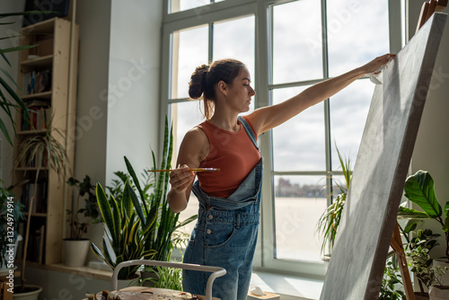 Thoughtful woman artist working on painting. Girl outstretched hand with brush reaches for canvas, making careful thoughtful strokes, creating masterpieces. Working process in art gallery, hobby. 