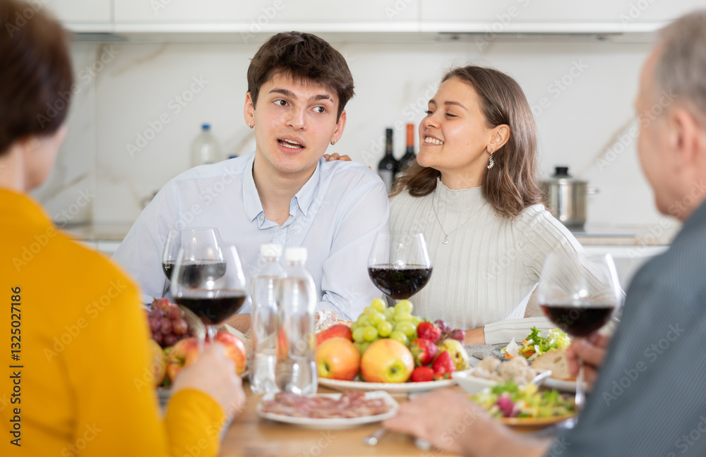 Cheerful young couple engaging in lively conversation with elderly parents at dinner table with wine and fruits in cozy home kitchen.. Cheerful young couple engaging in lively conversation with elderly parents at dinner table with wine and fruits in cozy home kitchen..