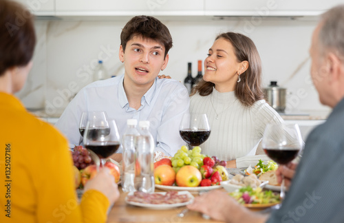 Wallpaper Mural Cheerful young couple engaging in lively conversation with elderly parents at dinner table with wine and fruits in cozy home kitchen.. Torontodigital.ca