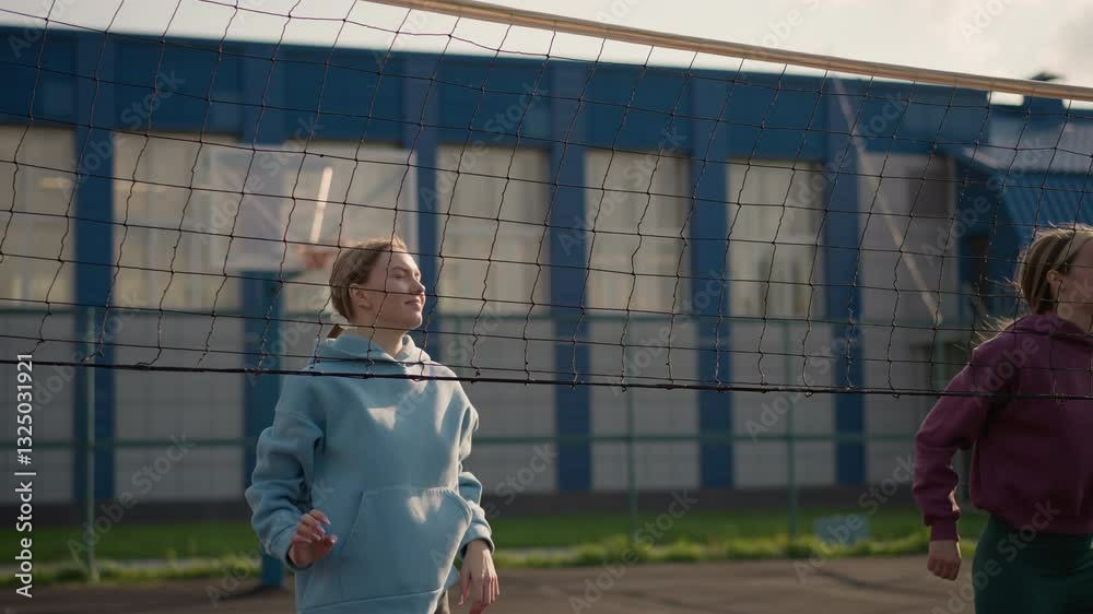 Beginners practicing volleyball outdoors with net in front, jumping up with hands open wide, background features a building and sports ground in bright sunlight