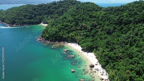Aerial view of Ilha das Couves located in Ubatuba, Brazil. People enjoy the beach and the sea water crashes against the rocks on the coast. Green trees are seen in the beautiful landscape.