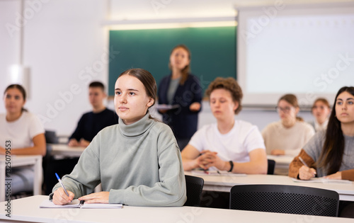 Group of school teenagers with pens and notebooks studying in classroom with teacher