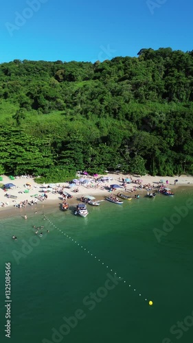 Aerial view of Ilha do Prumirim located in Ubatuba, Brazil. People enjoy the beach and the sea water crashes against the rocks on the coast. Green trees are seen in the beautiful landscape.